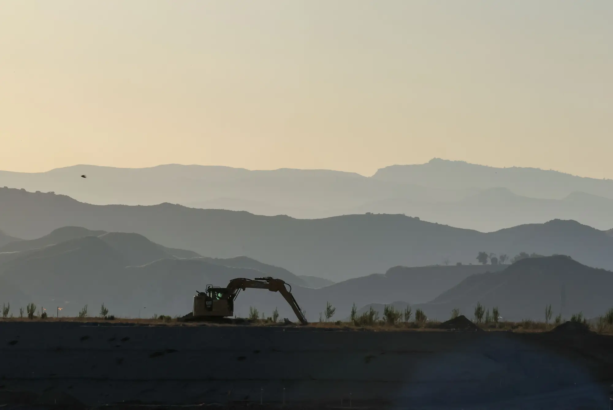 An excavator parked on a graded ridge at dusk, hills fading behind it
